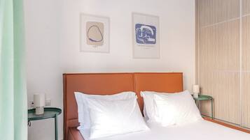 A hotel room with a bed, white pillows, a brown headboard, two framed artworks on the wall, and a green side table with a lamp.