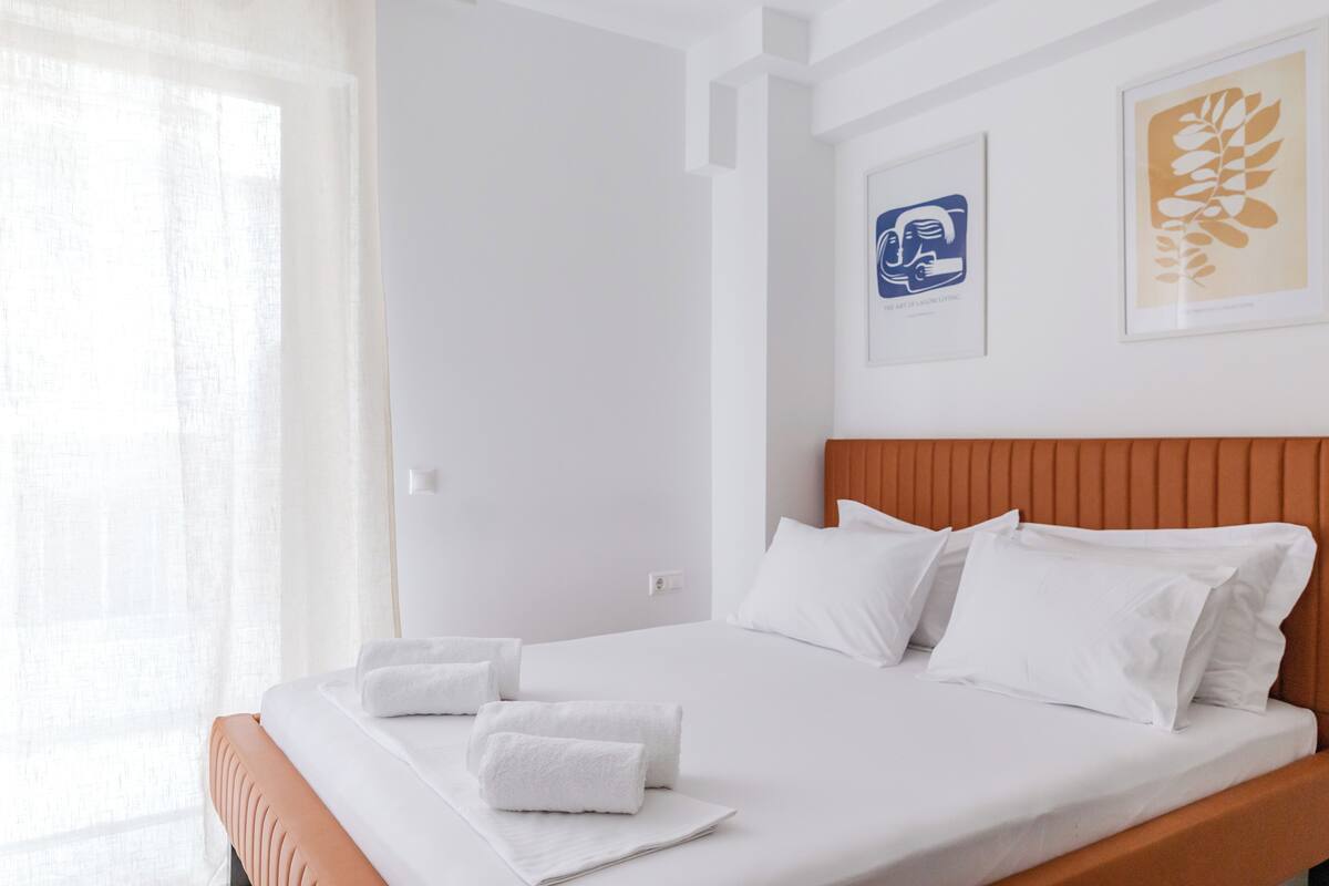A neatly made bed with white linens and pillows, a wooden headboard, and two framed artworks on the wall.