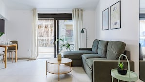 Moderni olohuone, jossa on sohva, pyöreä sohvapöytä ja pieni sivupöytä, jonka päällä on kasvi. Olohuoneessa on myös ruokailutila, jossa on pöytä ja tuolit, sekä suuri ikkuna, jossa on verhot.