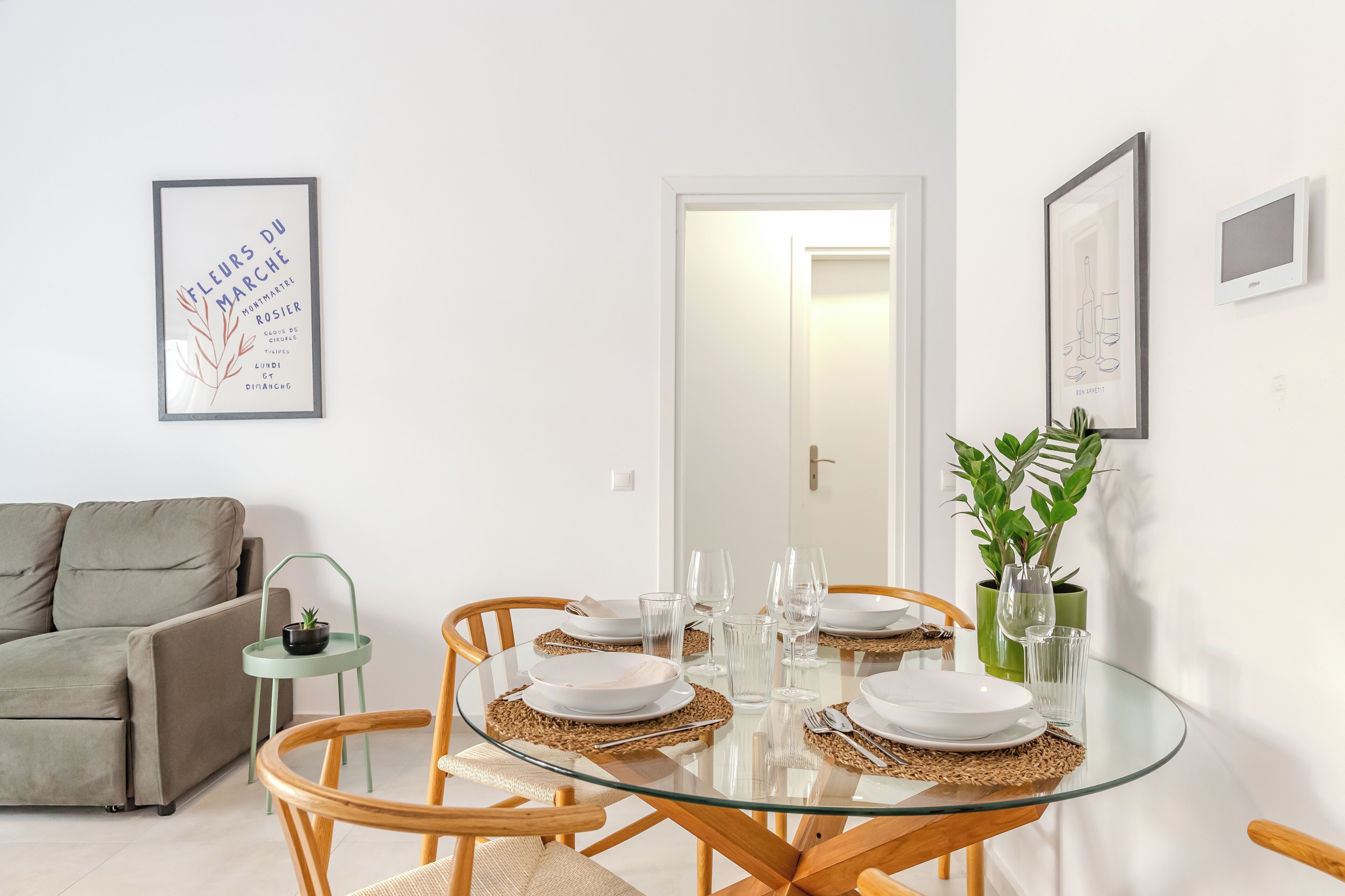 A dining area with a glass table, wooden chairs, and a sofa in the background.