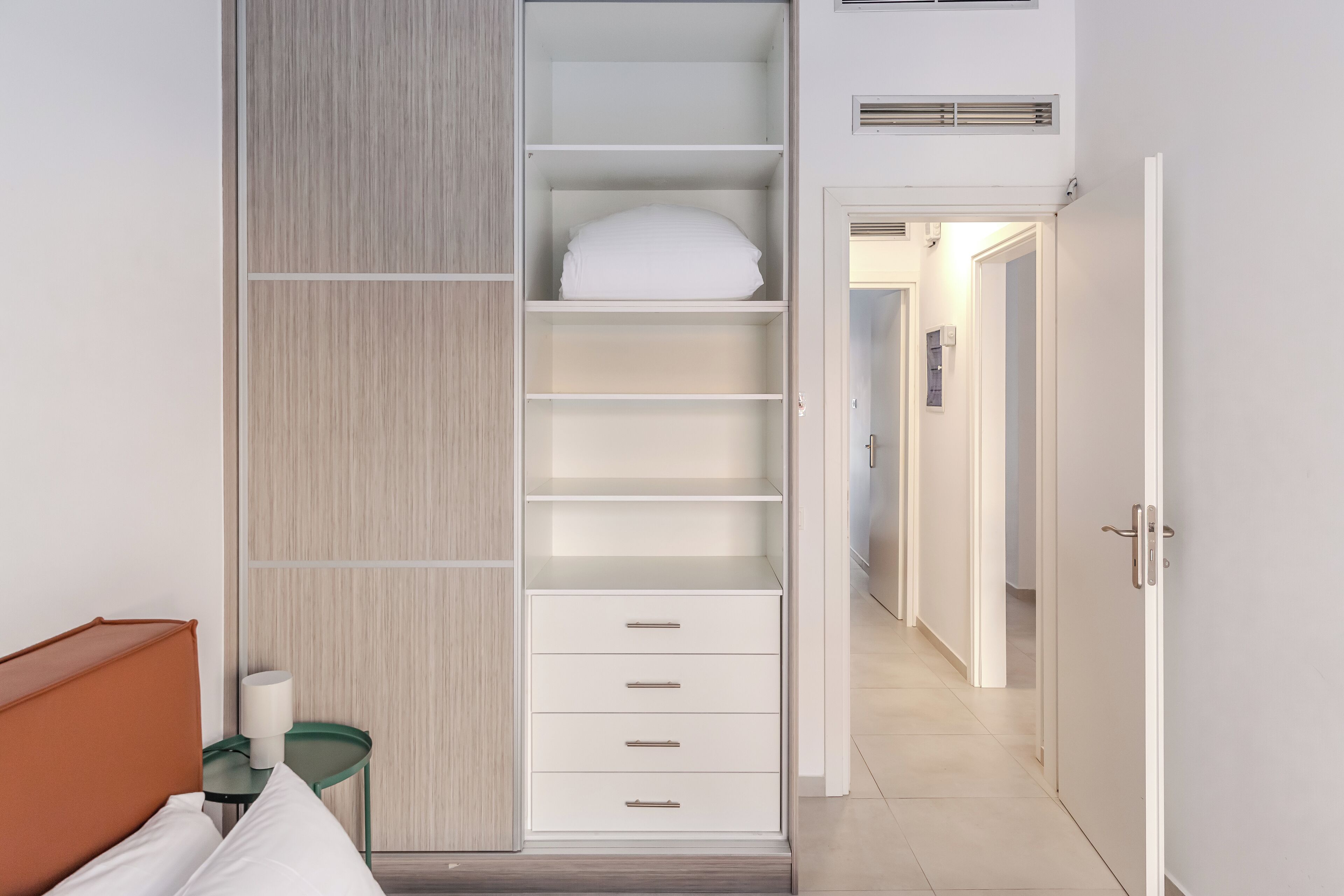 Ein Hotelzimmer mit einem Bett, einem Kleiderschrank mit mehreren Regalen und Schubladen, einem kleinen Tisch mit einer Lampe und einer Tür zu einem anderen Raum.