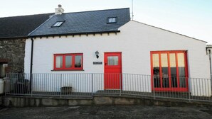 A white building with a red door and windows, a small porch, and a stone wall adjacent to it.