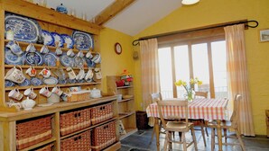 A dining area with a table set for two, wooden shelves with blue and white dishes, wicker baskets, and a window with curtains.