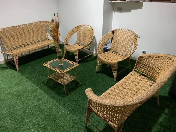 Una habitación con suelo de césped artificial verde, un sofá de mimbre, un sillón de mimbre, una mesita de noche de mimbre y un jarrón pequeño con plantas secas.