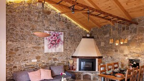 A cozy room with a stone wall, wooden ceiling beams, a fireplace, a sofa, a dining table with chairs, and hanging lights.