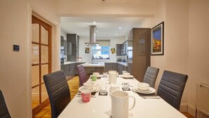 A dining area with a white table set for four, surrounded by black chairs. A kitchen is visible in the background with modern appliances. - The View Old Coastguard Cottage (Spanish Battery)