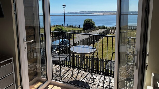 A balcony with a table and chairs, a view of the sea, and a distant shoreline. - The View Old Coastguard Cottage (Spanish Battery)
