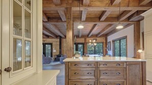 A spacious kitchen with a large island, wooden beams on the ceiling, and large windows providing natural light. - Chalet de l'aube | Spa (Sainte-Béatrix)