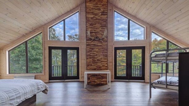 A spacious loft bedroom with a sloped ceiling, large windows, a stone fireplace, and a bunk bed. - Chalet de l'aube | Spa (Sainte-Béatrix)
