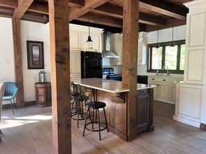A kitchen with a central island, bar stools, wooden beams, and a window with greenery outside. - Chalet de l'aube | Spa (Sainte-Béatrix)