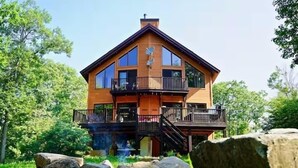 A two-story wooden house with a balcony on the upper floor, surrounded by trees and rocks. - Chalet de l'aube | Spa (Sainte-Béatrix)