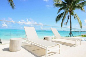 A beachside lounge with white lounge chairs and a concrete pillar, overlooking a clear blue sea and palm trees.
