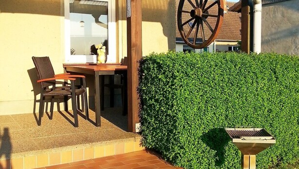 An outdoor patio with a table and chairs, a grill, and a hedge.