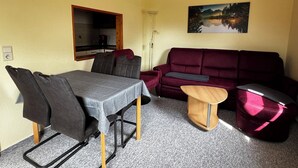 A compact living space with a dining table, chairs, a sofa, a small wooden side table, and a painting on the wall.