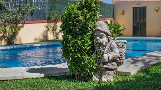 Une statue de pierre représentant un personnage barbu, placée au bord d’une piscine, avec un toit en tuiles et de la verdure en arrière-plan.