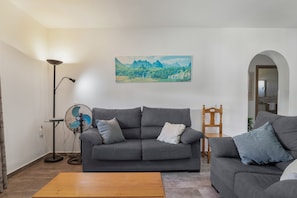 A living room with a grey sofa, a wooden coffee table, a standing fan, and a painting of mountains on the wall.
