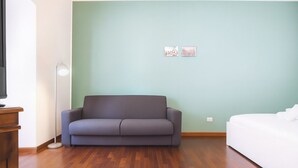 Una stanza con un divano grigio, un letto bianco, un pavimento in legno e una lampada da terra.