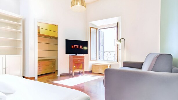 Una camera d'albergo moderna con un letto, un divano grigio, una televisione che mostra 'NETFLIX', un mobiletto in legno e una finestra con vista.