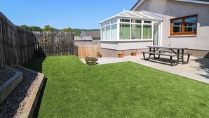 A backyard with a well-maintained lawn, a wooden fence, a wooden deck, and a house with a large window and a door. - Fircroft (Perth)