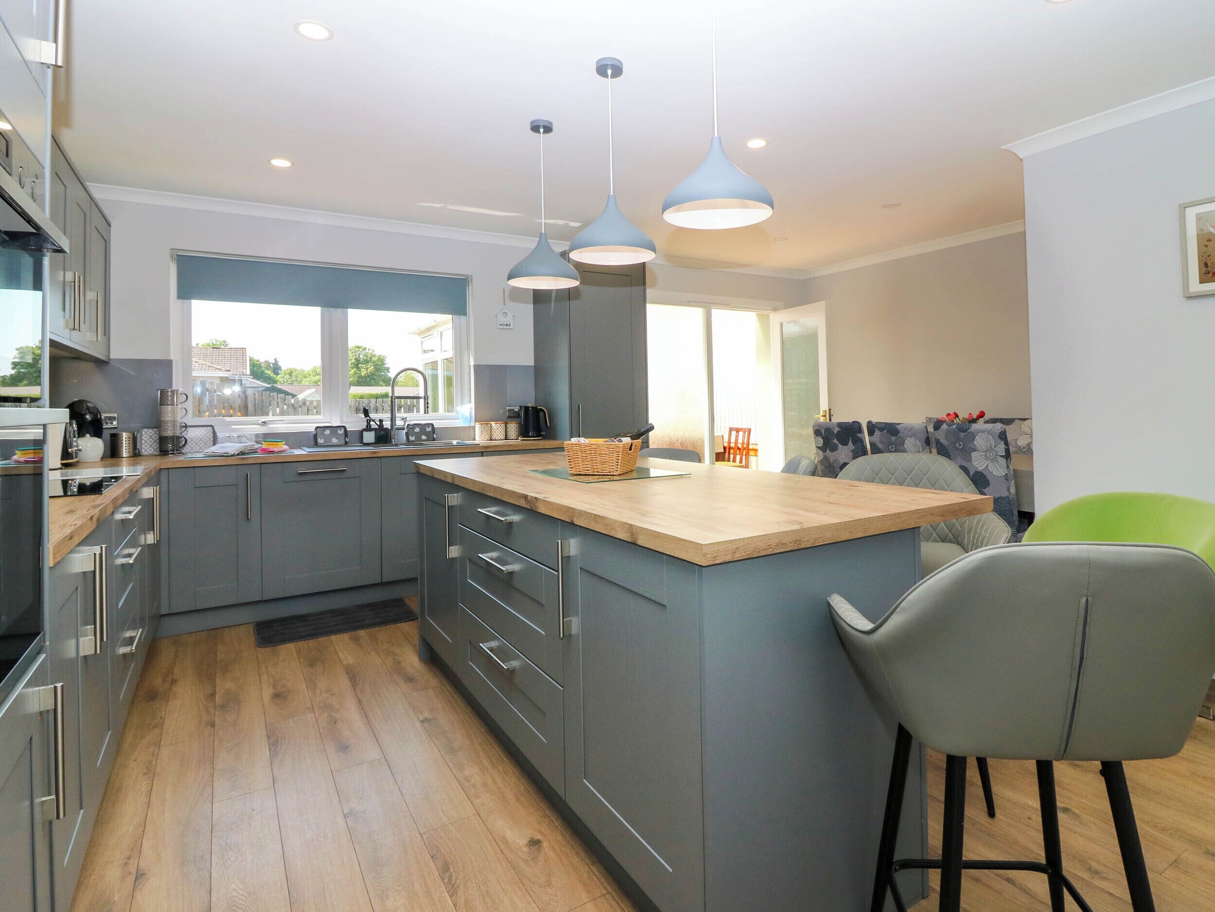 A modern kitchen with grey cabinets, wooden countertops, and a central island. There are hanging pendant lights, a window with a view, and a bar stool.