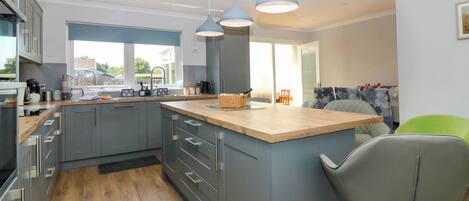 A modern kitchen with grey cabinets, wooden countertops, and a central island. There are hanging pendant lights, a window with a view, and a bar stool.