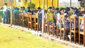 Un grupo de personas sentadas en mesas de madera bajo un techo de paja, con un edificio al fondo que dice 'BUNGALOWS'.