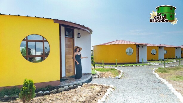 Un edificio amarillo con un ventanal circular y una puerta, rodeado por un camino de grava y pequeñas plantas.