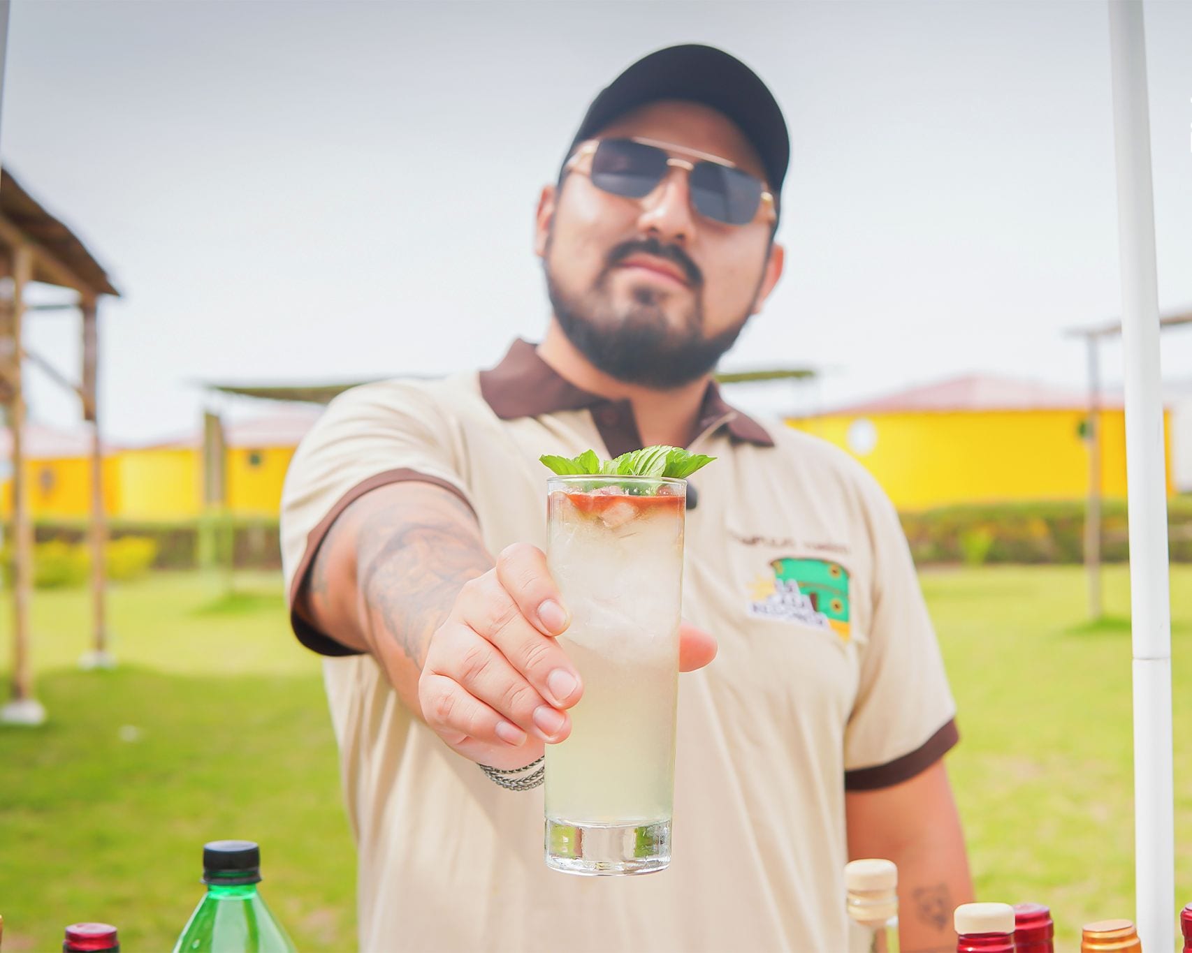 A person holding a tall glass with a drink, wearing a shirt with a logo, outdoors with buildings in the background.