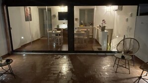 Un espace extérieur pavé, exposé aux intempéries, avec une porte vitrée donnant sur un espace intérieur comprenant une table à manger, des chaises et un coin cuisine.