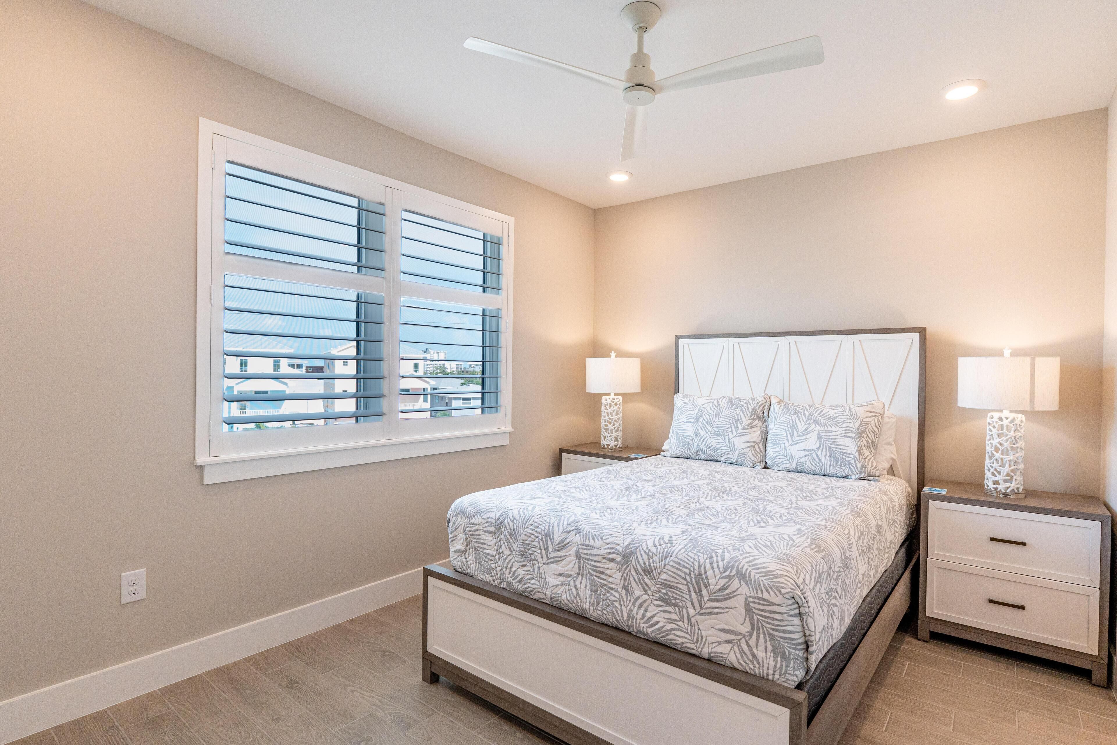 A bedroom with a bed, two bedside tables with lamps, a ceiling fan, and a window with blinds.