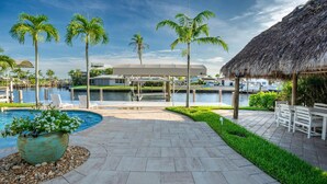 A resort pool area with a thatched-roof structure, palm trees, and a water body. - High Tide Hideaway (Fort Myers Beach)