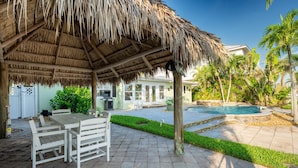 A covered patio with a thatched roof, a pool, and a dining area with white furniture. - High Tide Hideaway (Fort Myers Beach)