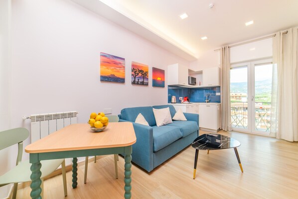 A modern living room with a blue sofa, a wooden dining table with a bowl of lemons, and a small black coffee table. There are three paintings on the wall depicting sunsets.