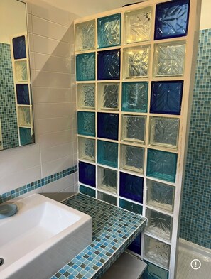 Ein Badezimmer mit einer Badewanne, einem Spiegel und einer Wand mit quadratischen Glasfliesen in verschiedenen Blautönen und Grüntönen.