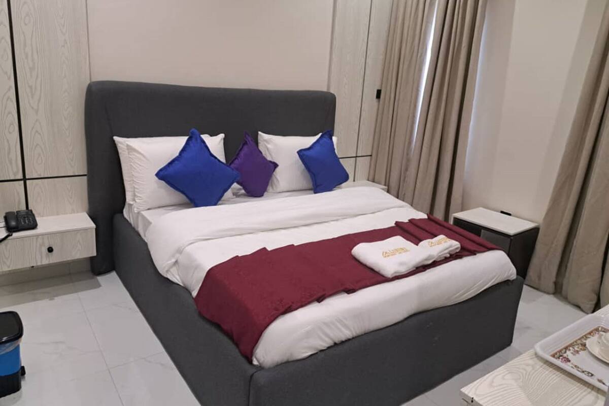 Een net opgemaakt bed met wit en bordeauxrood beddengoed, een grijs hoofdbord en blauw-paarse kussens. Er staat een klein nachtkastje met een lamp en een telefoon, en een blauwe prullenbak op de vloer.