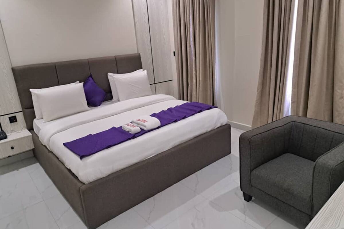 Een hotelkamer met een bed, nachtkastje, stoel en raam met gordijnen.