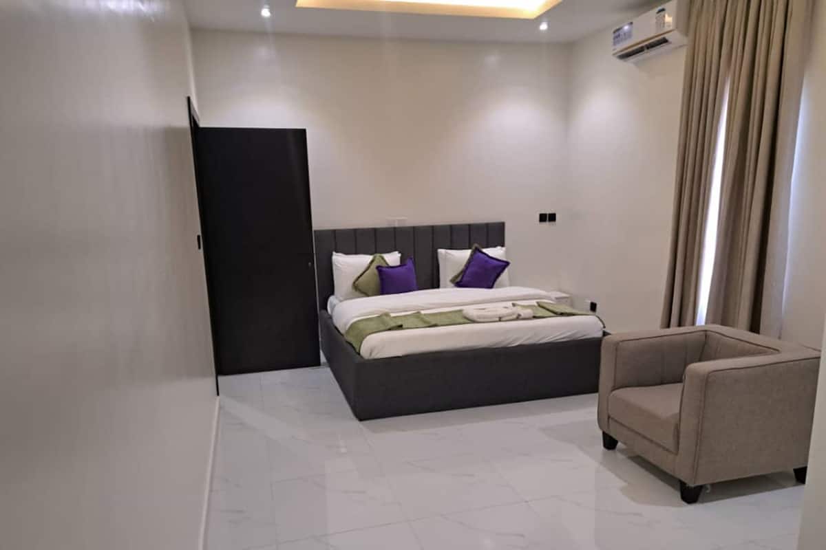 Een moderne hotelkamer met een groot bed, een stoel en een deur naar een andere kamer.
