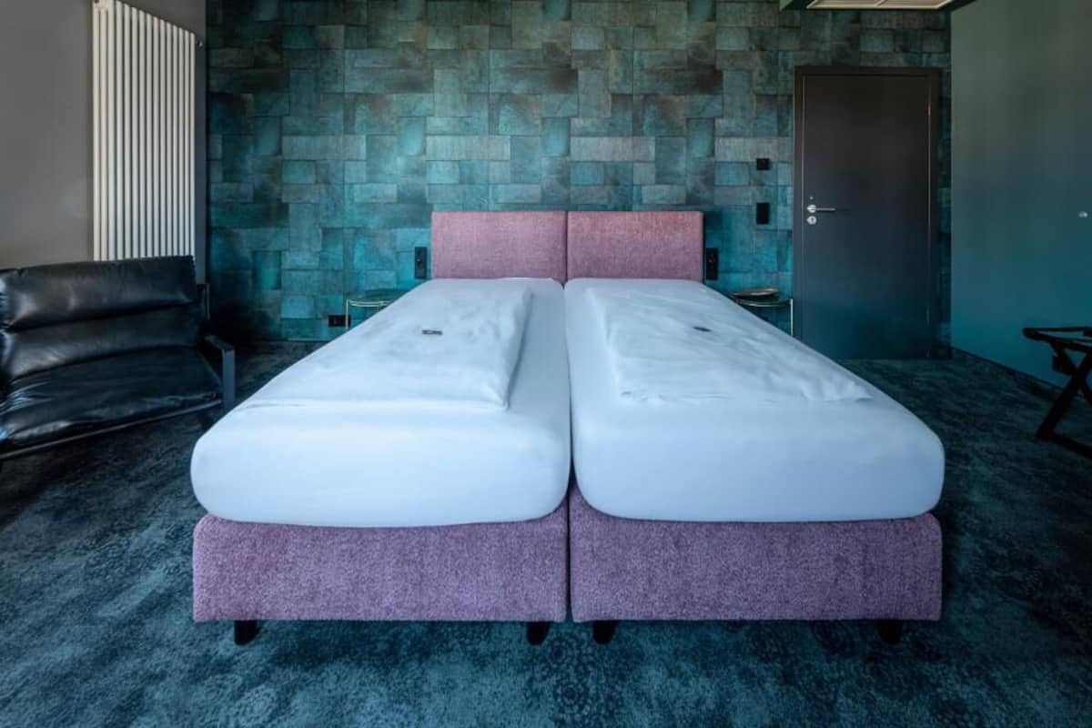 Een tweepersoonsbed met een roze hoofdbord in een kamer met een muur met steenmotief en een donker vloerkleed.