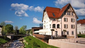 Ein traditionelles Fachwerkhäuschen mit rotem Dach, einem Fluss im Vordergrund und blauem, wolkenlosem Himmel.
