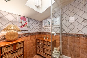 Une salle de bain avec un mur carrelé, une lampe en osier, des paniers en osier, une douche et une affiche.