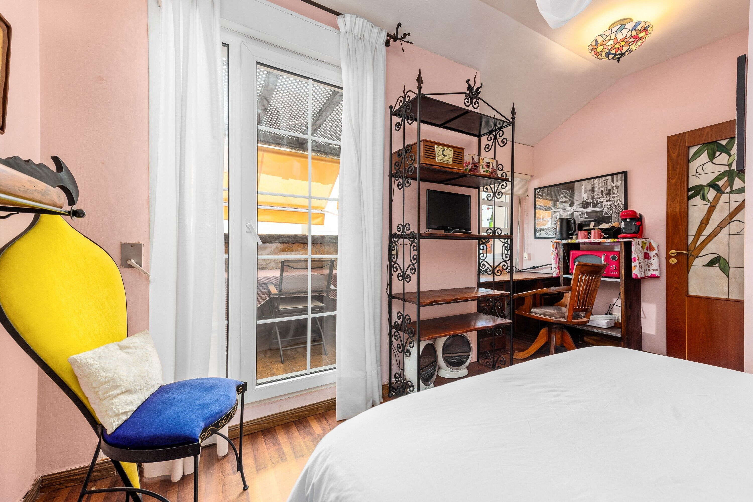 Ein Schlafzimmer mit einem Bett, einem Sessel, einem Schreibtisch mit Fernseher und einem Fenster mit Vorhängen.