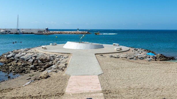 Une plage avec une structure circulaire en béton, un sentier menant à l’eau et une côte rocheuse.
