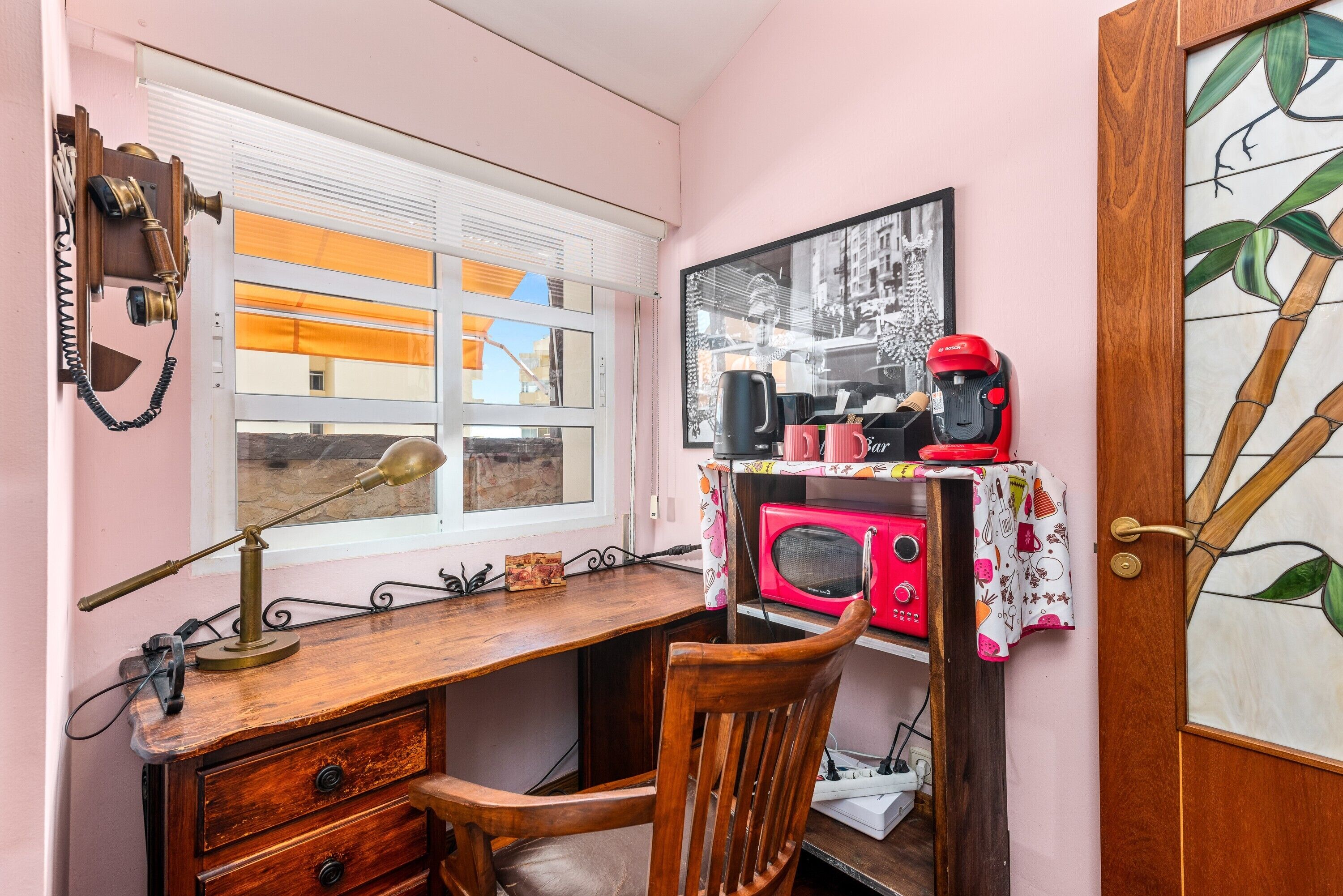 Ein Zimmer mit einem Holztisch, einem Stuhl, einem Fenster mit Jalousien, einem gerahmten Bild an der Wand und einer Tür mit Glasfenster.