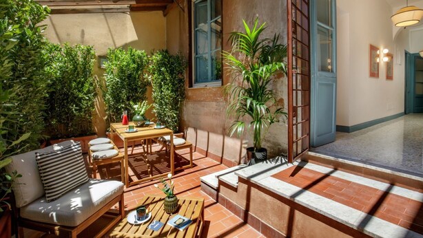 Eine sonnendurchflutete Terrasse mit Holztisch und -stühlen, umgeben von üppigem Grün und einem großen Fenster.