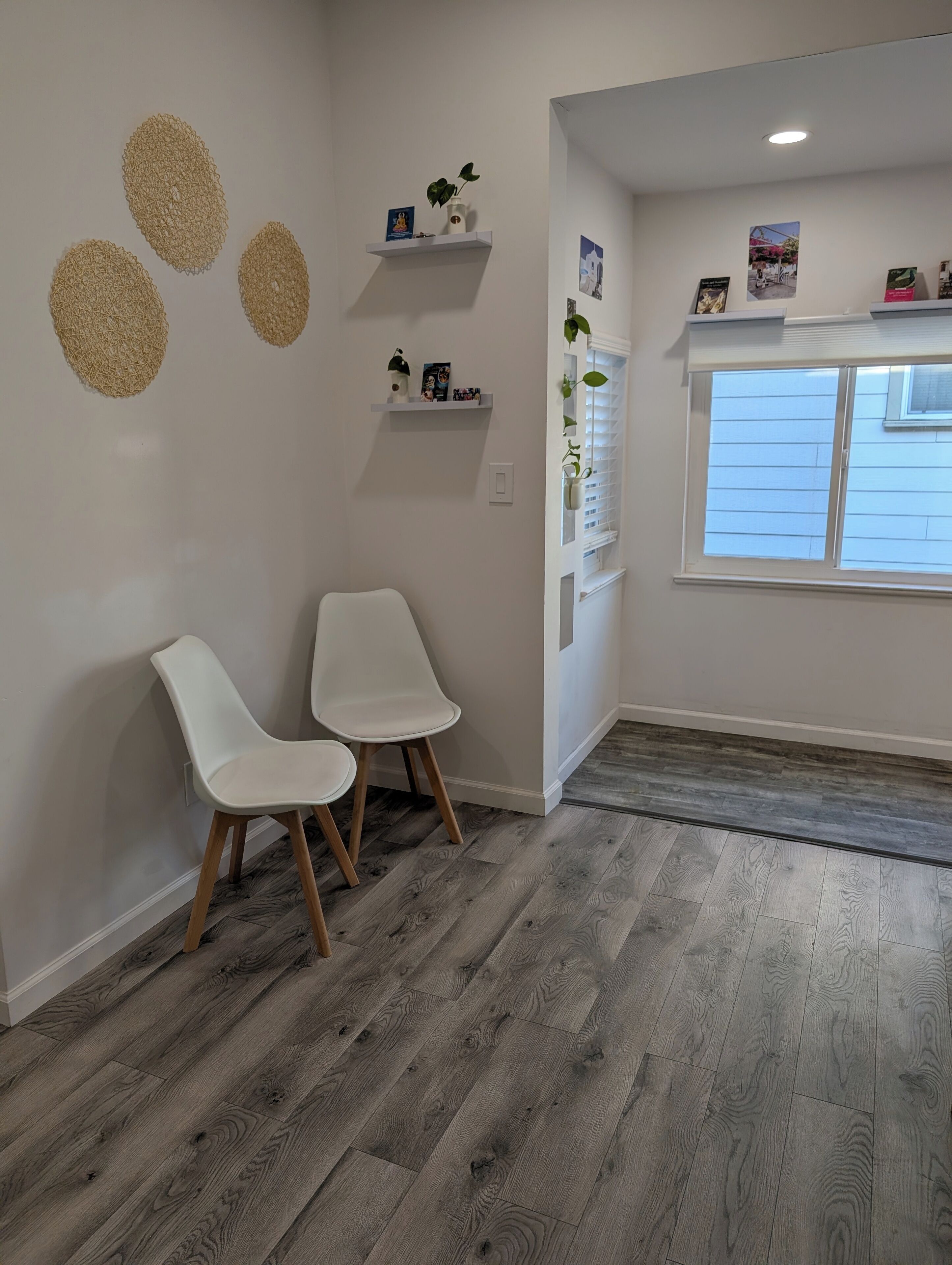 A room with two white chairs, a wooden floor, a white wall with decorative circles, a shelf with small items, and a window with blinds.