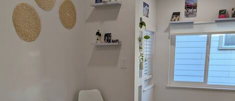A room with two white chairs, a wooden floor, a white wall with decorative circles, a shelf with small items, and a window with blinds.
