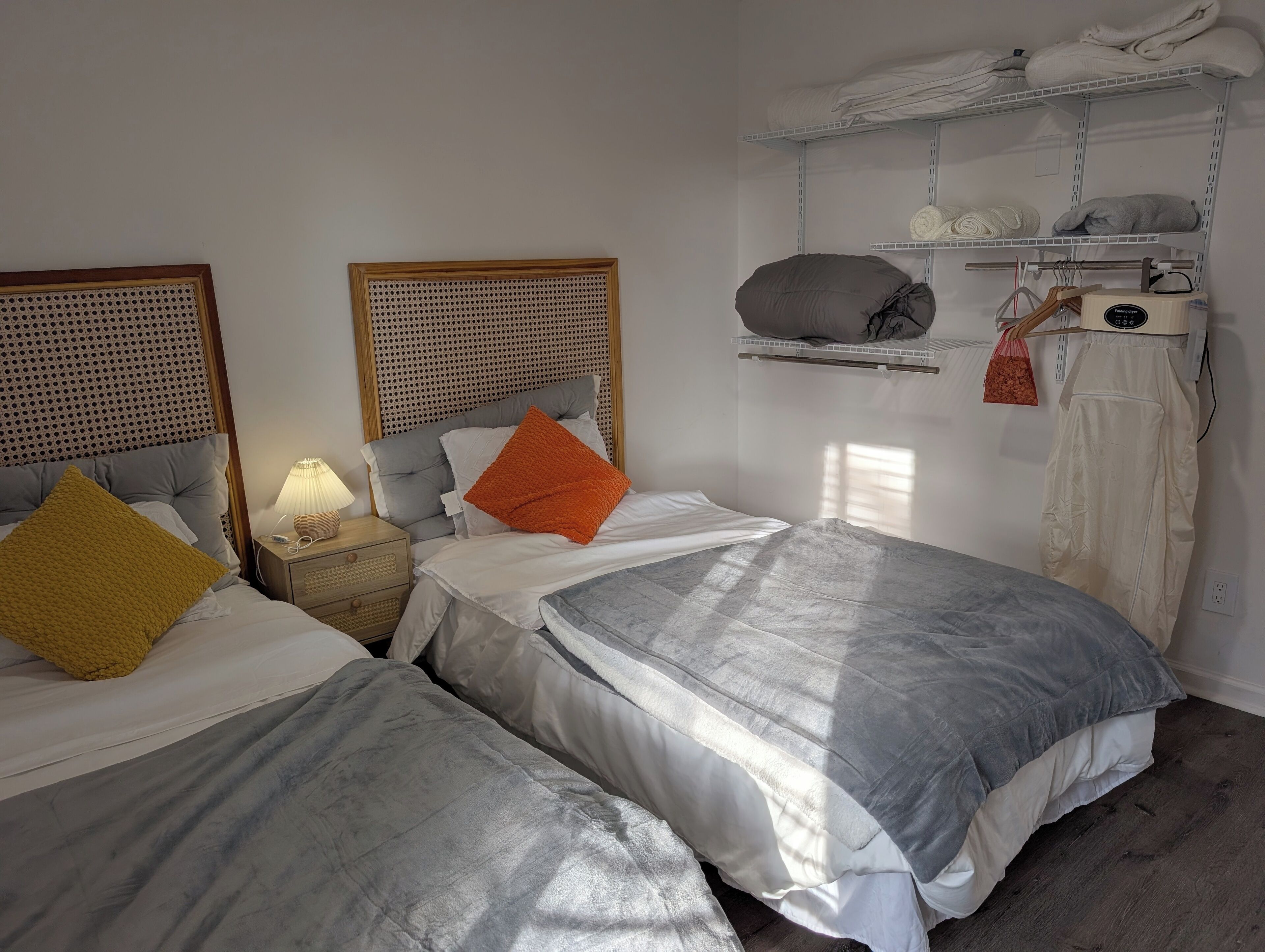A bedroom with two beds, a nightstand with a lamp, a shelf with towels and a bag, and a hanging laundry rack.