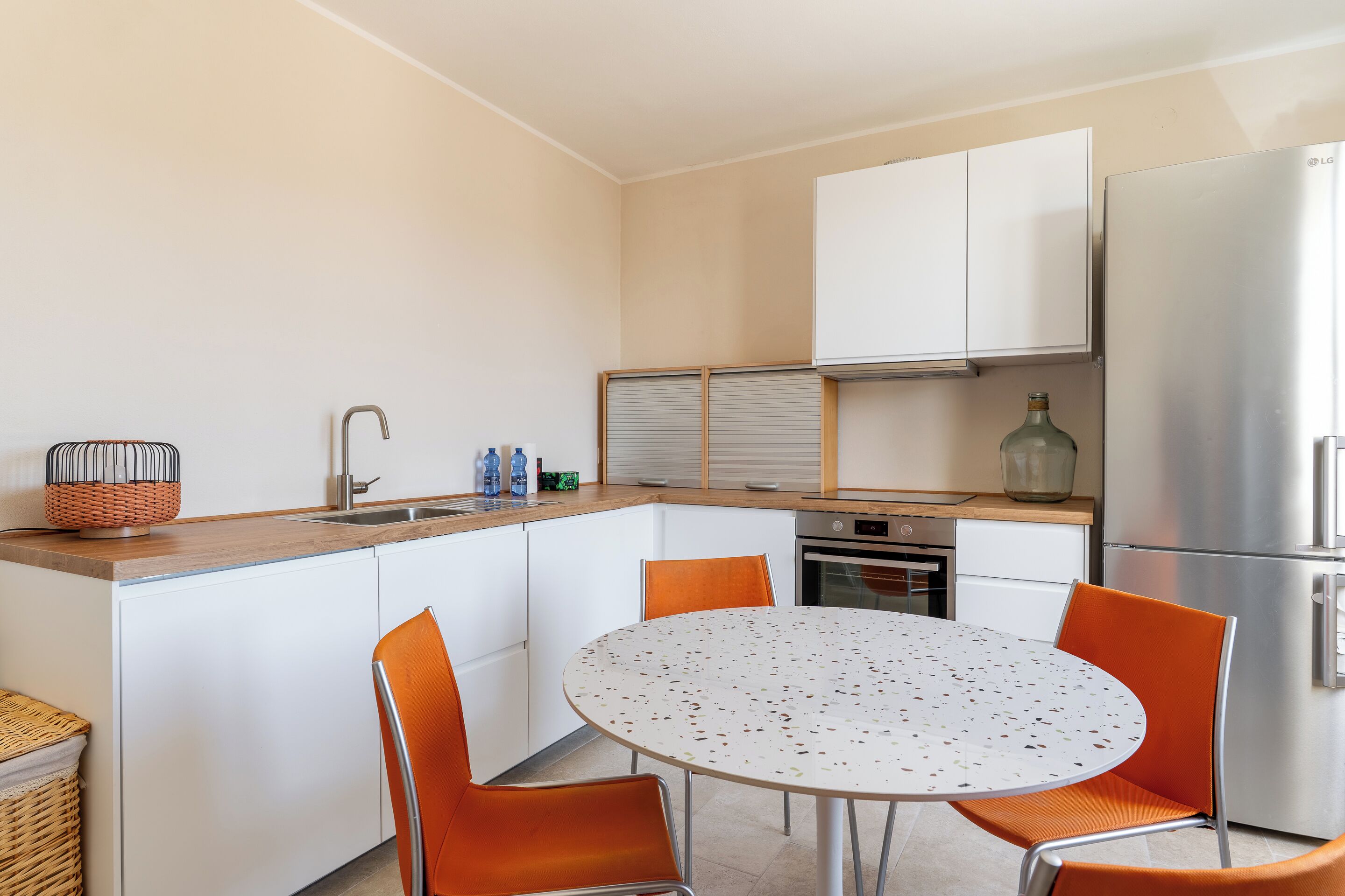 A compact kitchen with white cabinets, a stainless steel refrigerator, and a round table with orange chairs.