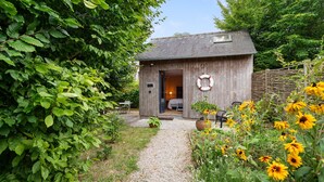 Una baita in legno con un vialetto sterrato che conduce all'ingresso, circondata da vegetazione e fiori gialli.
