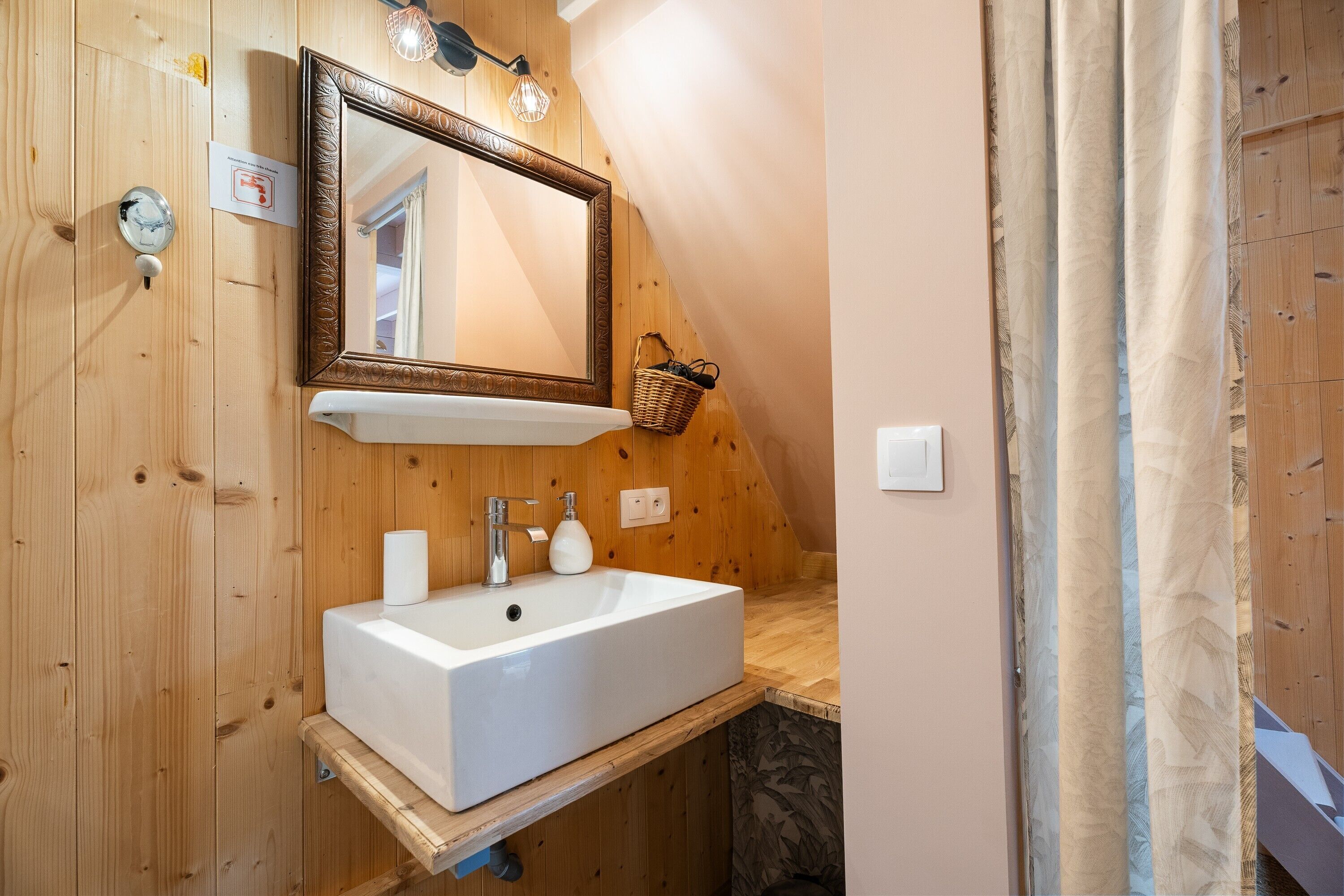 Une salle de bain aux murs lambrissés, avec un lavabo blanc, un miroir et un distributeur de serviettes.